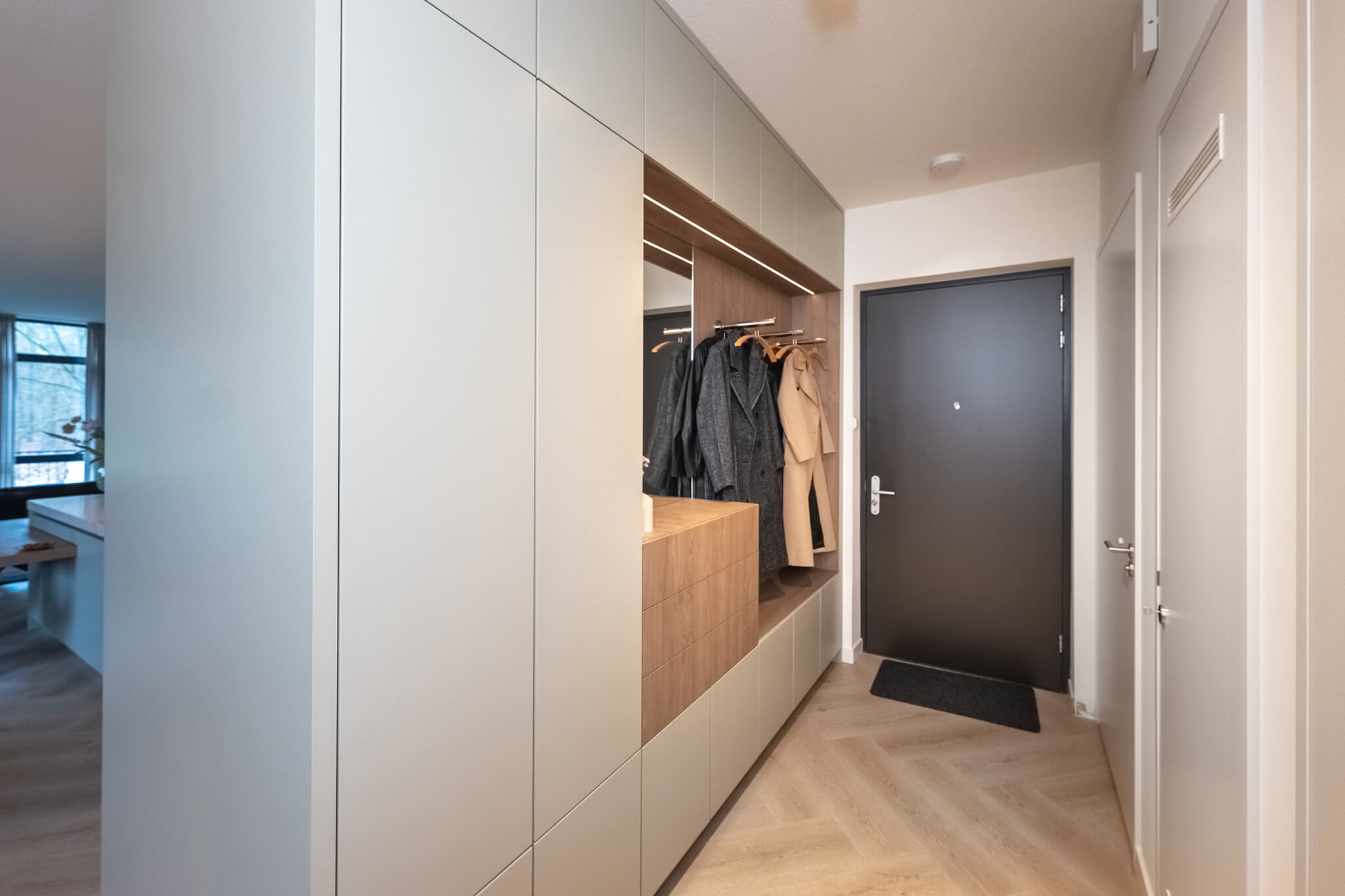 Een hoge kastenwand in de gang met vakken voor jassen en schoenen uitgevoerd in een combinatie van grijs en warm hout.